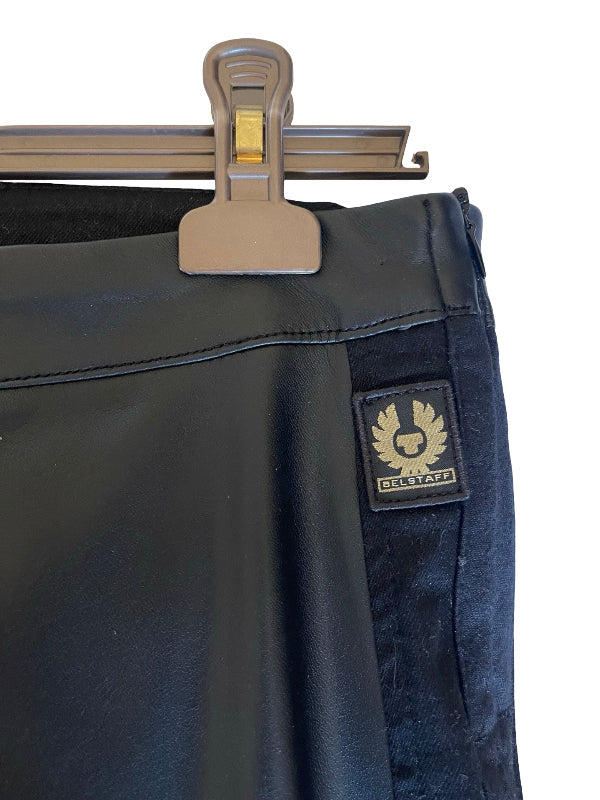 Close-up of black denim jeans hanging from a metal hanger, showing a black brand patch with a gold eagle logo and the word 'Belstaff' on the side seam.