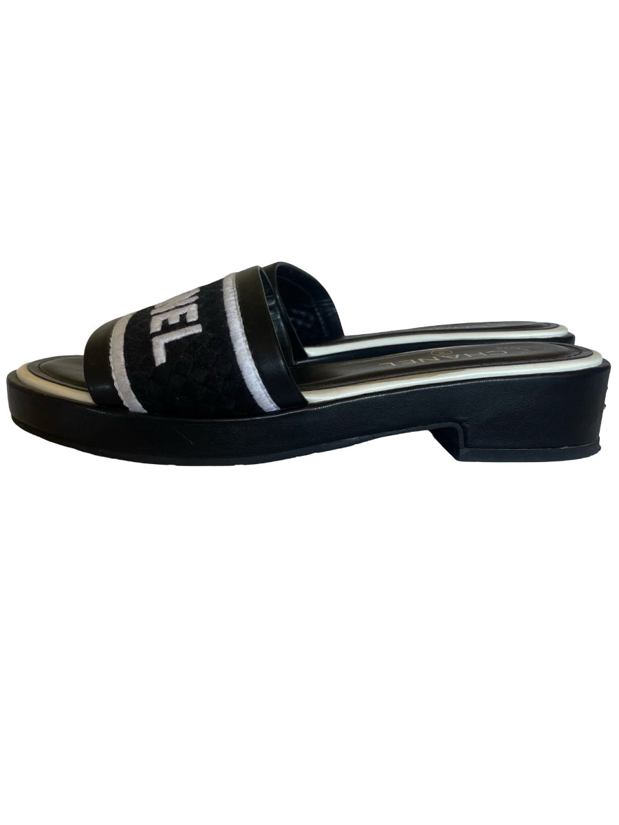A black platform slide sandal with a wide strap featuring word in white lettering and two diagonal white stripes, shown from a side profile on a white background.