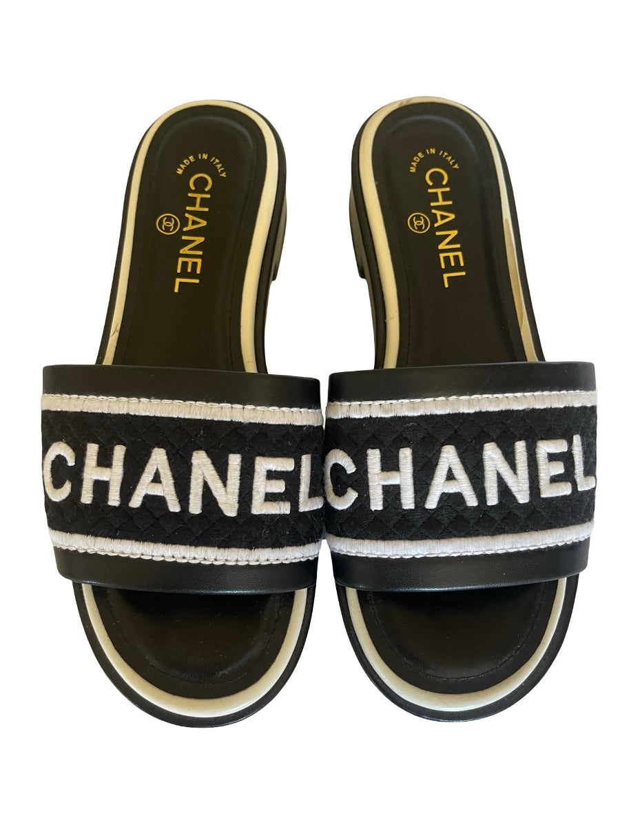 A pair of Chanel style slide sandals with black woven straps featuring the word   CHANEL in white capital letters, bordered by white stripes, on a white background.