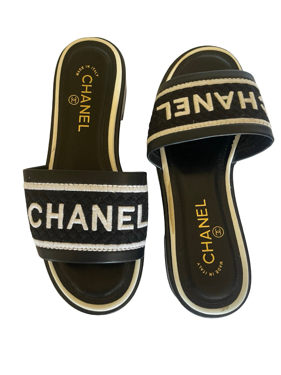 A pair of  slide sandals with black straps and soles, featuring white and gold branding details, set against a plain white background.