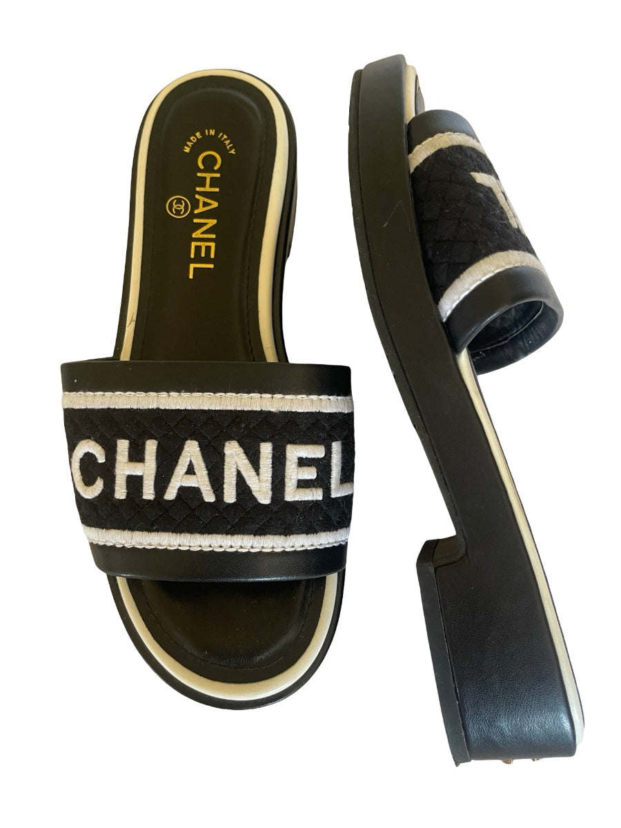 A pair of slide sandals shown from the top and side views on a plain white background. The sandals feature a black woven strap with a prominent white embroidered logo and double stripe accents, set on a black platform sole with cream-colored trim.