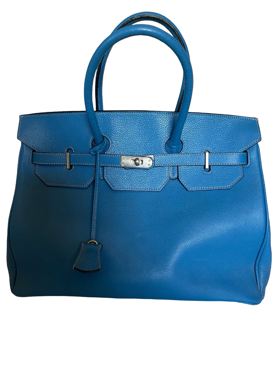 A blue leather handbag with double top handles, a front flap, silver-tone hardware, and a hanging clochette, photographed against a plain white background.