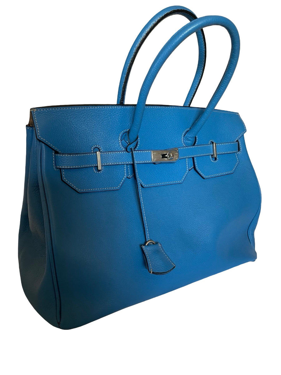 A medium blue leather handbag with silver-tone hardware, featuring dual top handles, a front flap with strap details, and a hanging clochette, shown against a plain white background.
