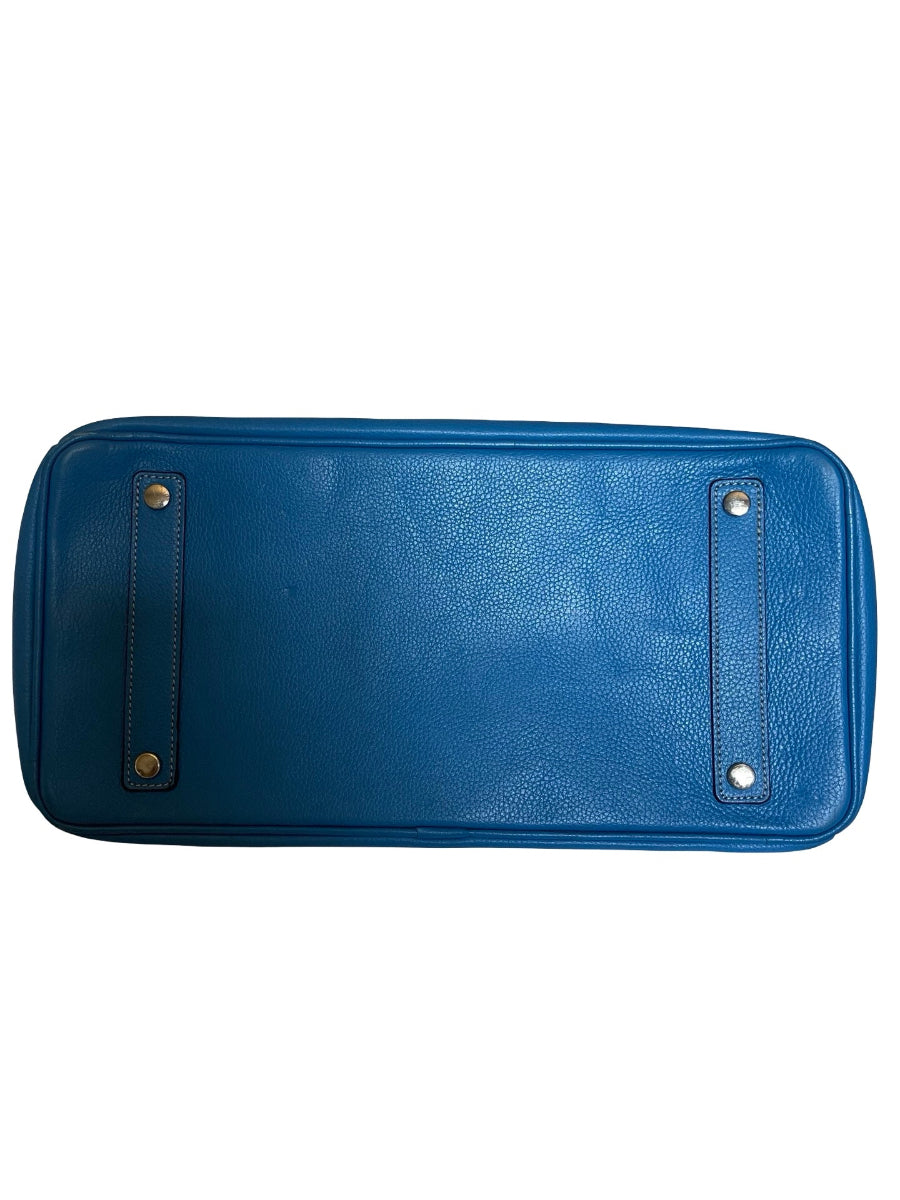 A blue pebbled leather handbag shown from the bottom, featuring two vertical strap details with gold-tone and silver-tone rivets, on a plain white background.