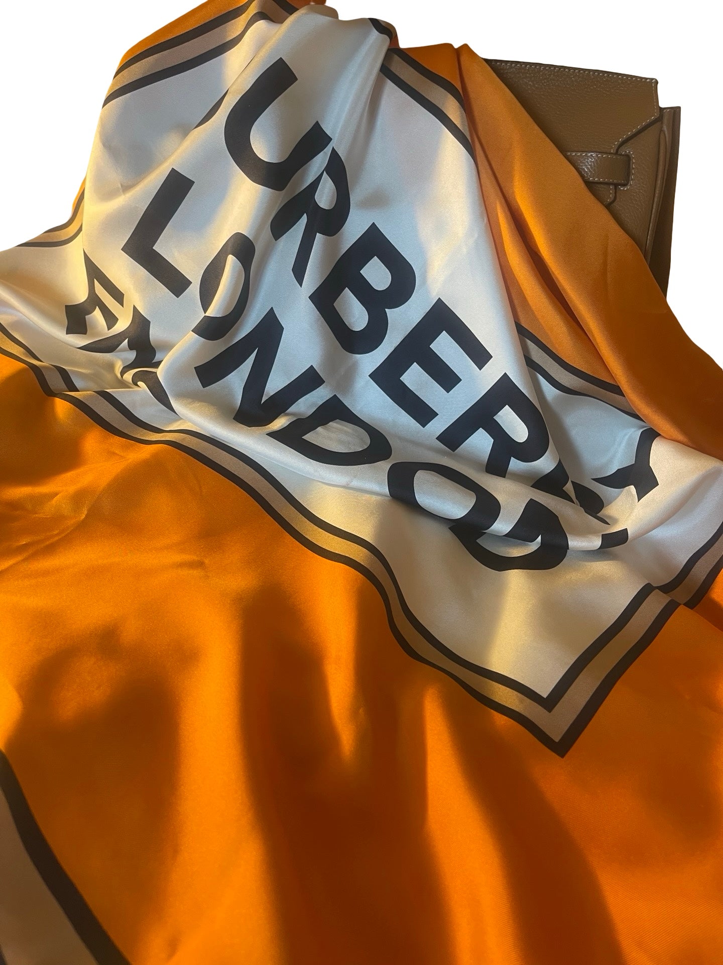 An orange and white silk scarf with black border stripes and the text 'BURBERY LONDON' printed in bold black letters, draped over a brown leather clutch bag.