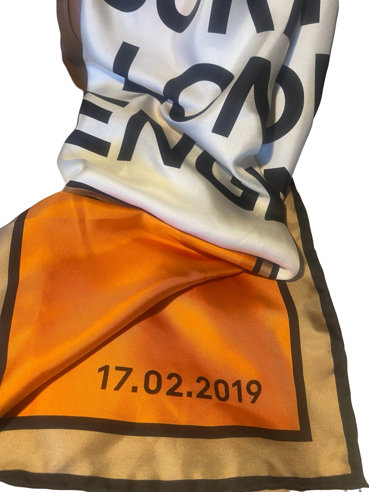 A close-up of a printed scarf with a white top section featuring large black letters, an orange lower section with the date '17.02.