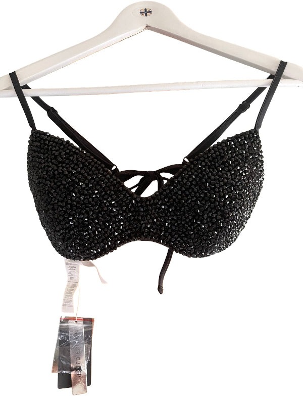 A black beaded bralette with thin shoulder straps and a front keyhole tie, hanging on a white wooden hanger against a white background, with retail tags attached below.