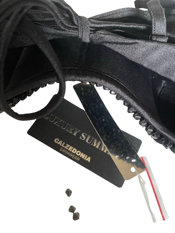 Close-up of a black satin swimwear top or bikini detail with lace-up straps, a black brand hangtag reading 'LUXURY SUMMER CALZEDONIA SWIMWEAR,' and a small plastic bag on a white background.
