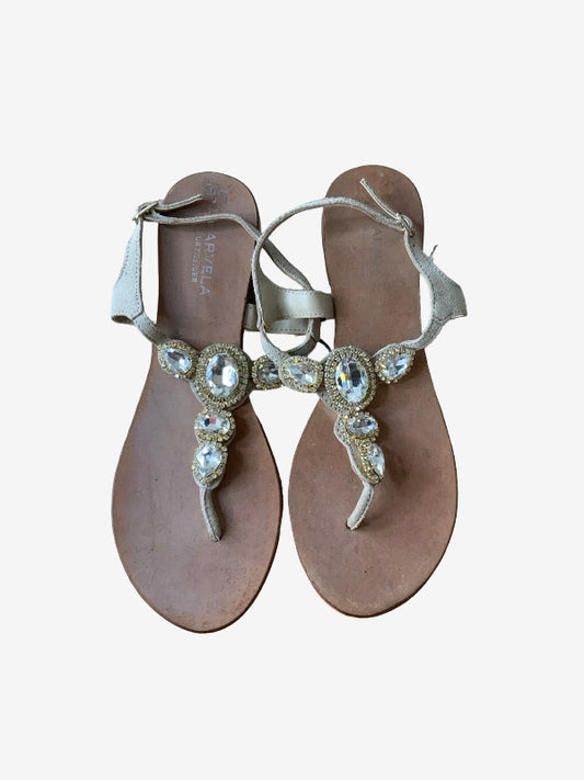 A pair of women's flat sandals with metallic silver straps adorned with large clear rhinestones and gold-toned detailing, set against a plain light gray background.