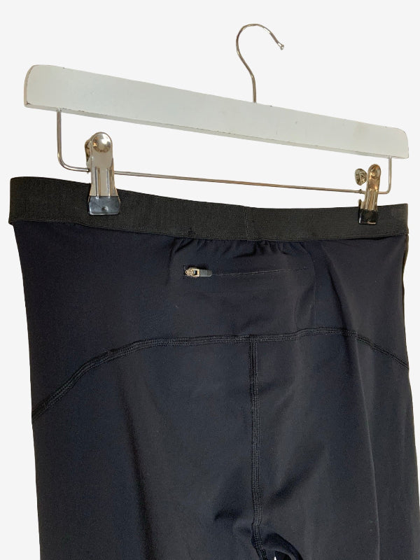 Close-up of the back waistband and pocket area of black athletic pants hanging from a white hanger with metal clips against a light gray background.