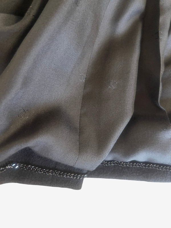 Close-up of a black silk fabric garment lining, showing a textured satin-like sheen, a fine chain trim along the hem, to weight the jacket and a white background at the bottom.