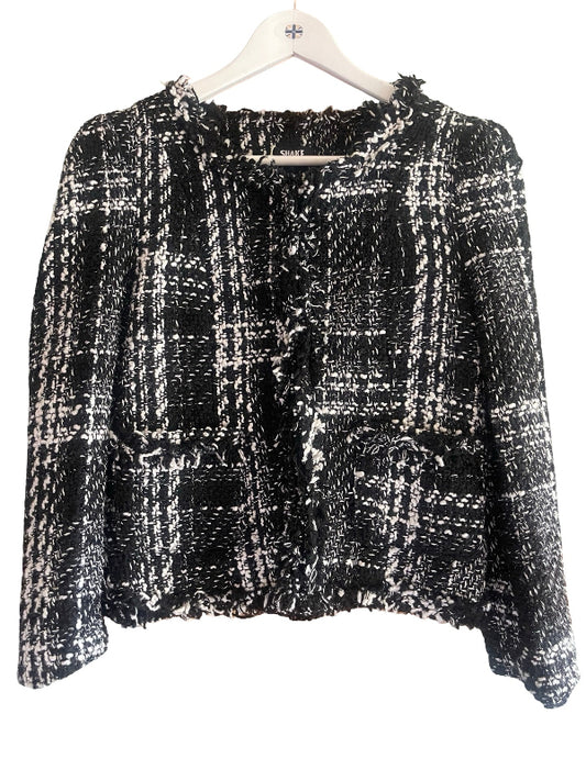 A black and white tweed plaid cropped jacket with long sleeves and a ruffled front, hanging on a white wooden hanger against a white background.