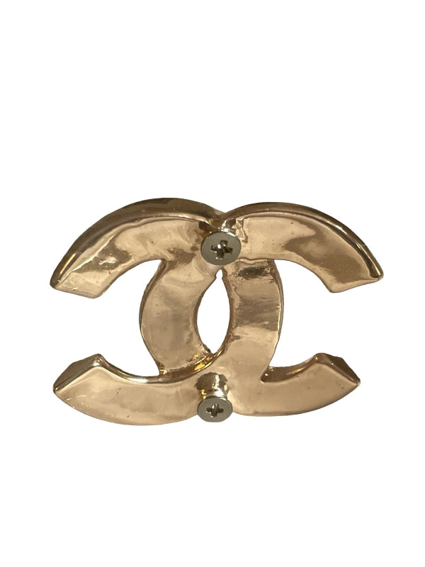 A gold-tone metal interlocking CC logo pin, characteristic of Chanel, with two small silver screws at the center of the interlocking design, shown on a plain white background.
