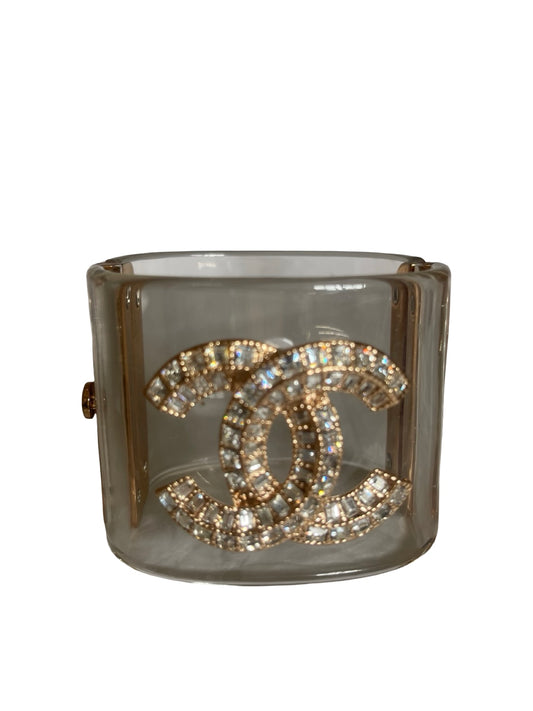 A Chanel cuff bracelet made of clear Perspex translucent with a large gold-tone interlocking CC logo paved with clear crystals, shown on a plain white background.