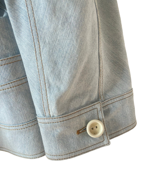 Close-up detail of a light blue denim jacket sleeve showing the cuff, contrast stitching, and a single cream-colored button on a white background.