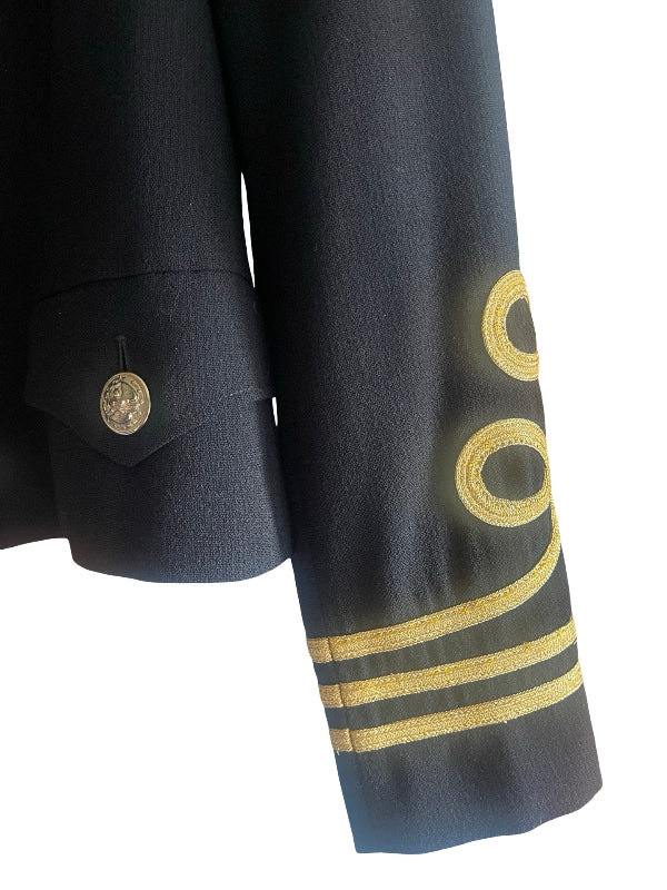 Close-up of a black uniform jacket sleeve with gold embroidered epaulettes and stripes, alongside a matching cuff tab with a gold button, on a white background.