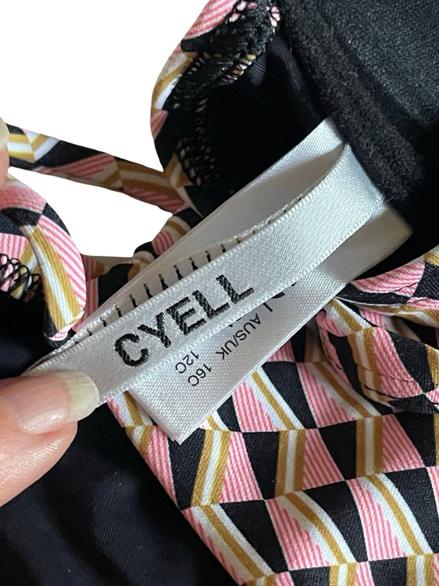 Close-up of a CYELL garment label attached to a black, pink, white, and gold striped patterned fabric, held by a finger.