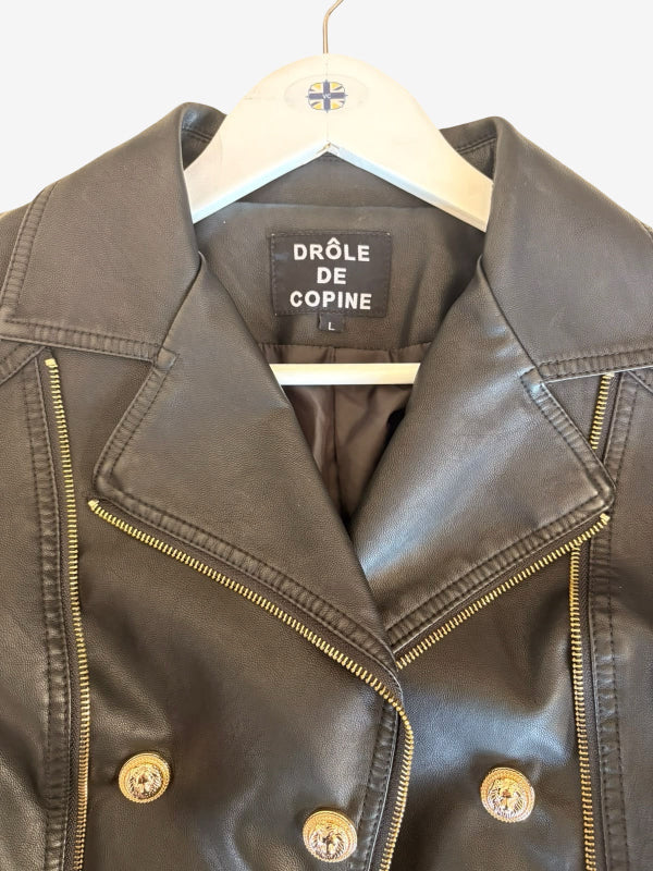 A close-up of a black faux  leather jacket hanging on a white wooden hanger. The jacket features asymmetrical gold-tone zippers, decorative gold-tone buttons, and a black brand label inside the collar that reads 'DRØLE DE COPINE'.