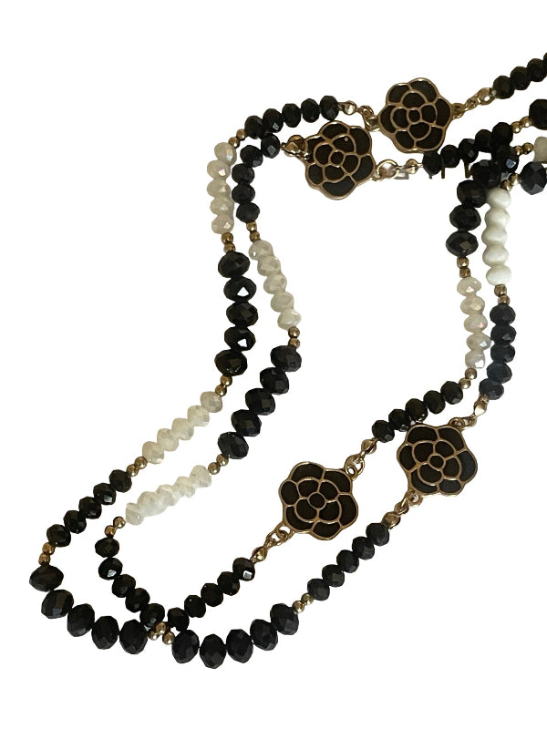 A multi-strand beaded necklace featuring black faceted beads, white pearl-like beads, and gold-tone floral medallions with black enamel detailing, displayed on a clean white background.