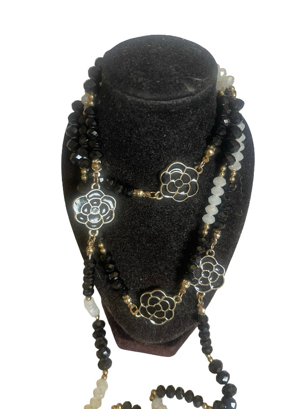 A multi-strand beaded necklace displayed on a black velvet jewelry bust. The necklace features black faceted beads, white opaque beads, gold-toned chain links, and silver-toned metal flower-shaped charms.