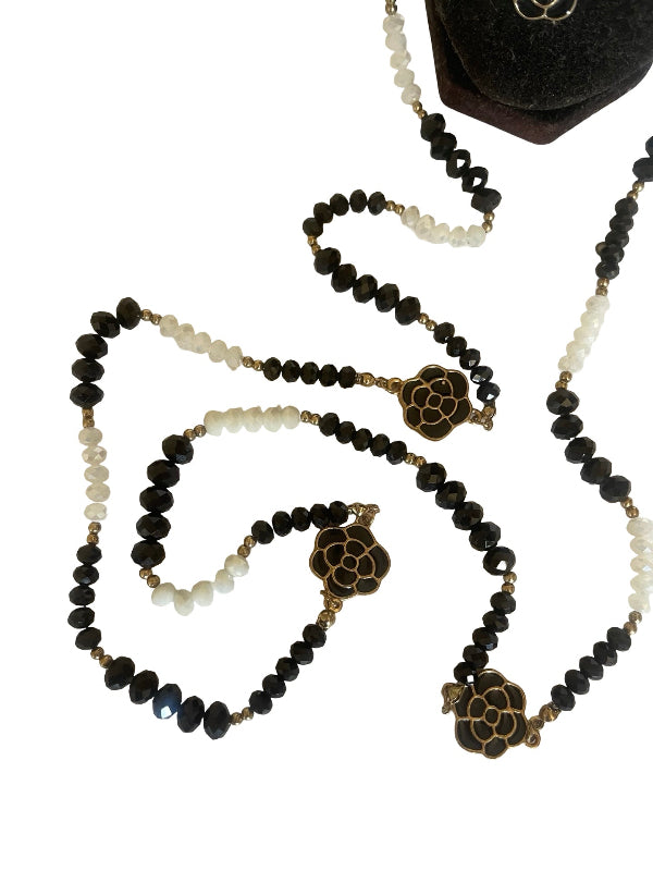 A beaded necklace with black faceted beads, white beads, and gold-toned floral charms, displayed on a white background with a black velvet jewelry stand partially visible at the top.