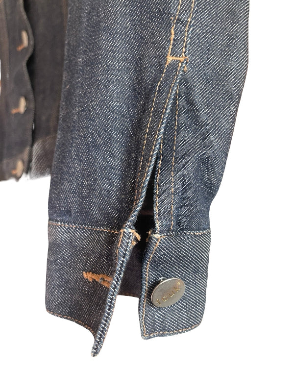 Close-up of a dark indigo denim jacket sleeve showing the buttoned cuff, contrast tan stitching, and a distressed area on the fabric, set against a plain white background.