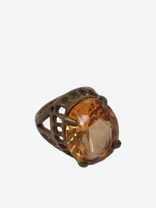 A ring with a large oval faceted amber-orange gemstone in a dark bronze-toned metal setting with openwork details, shown on a plain light gray background.