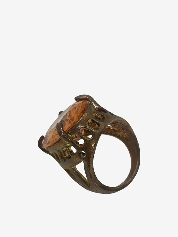 An ornate vintage-style ring with a large amber-brown faceted gemstone and a textured bronze-toned metal band, shown on a plain light gray background.