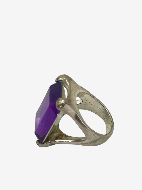 A silver-toned ring featuring a large, faceted purple rectangular gemstone, shown against a plain light gray background.