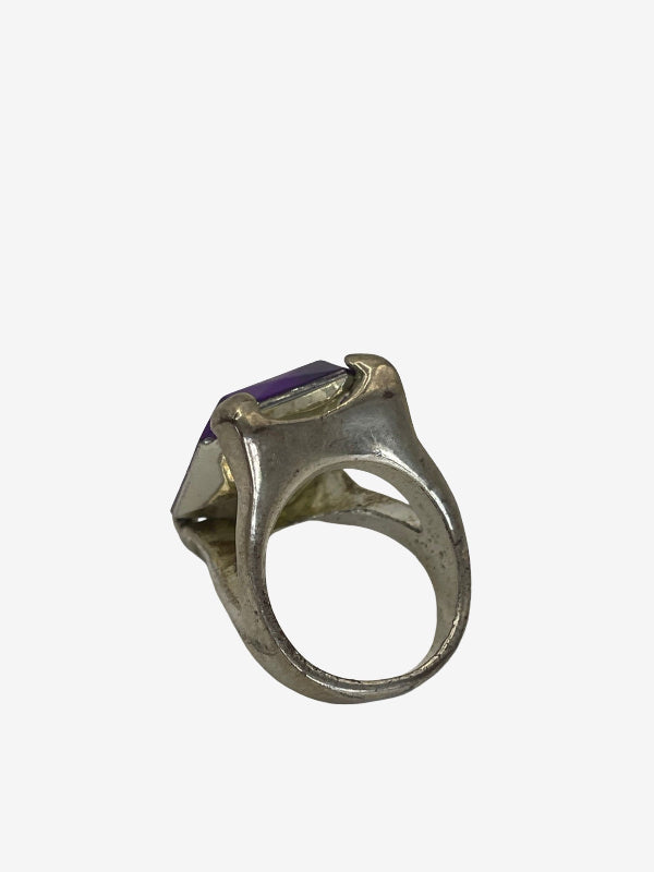 A silver-toned ring featuring a large, faceted rectangular purple gemstone, shown from an angled top-down view against a plain light gray background.