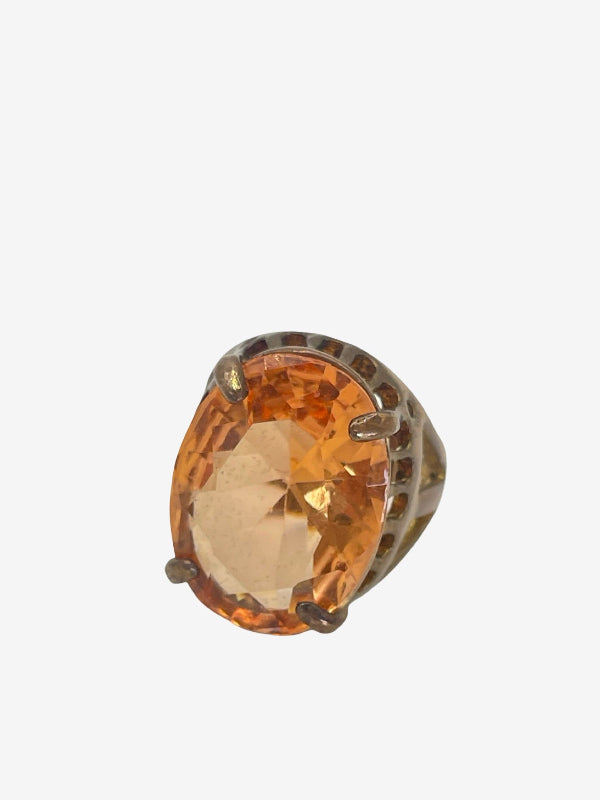 A single ring featuring a large, faceted oval orange gemstone in a silver-toned setting, photographed against a solid light gray background.