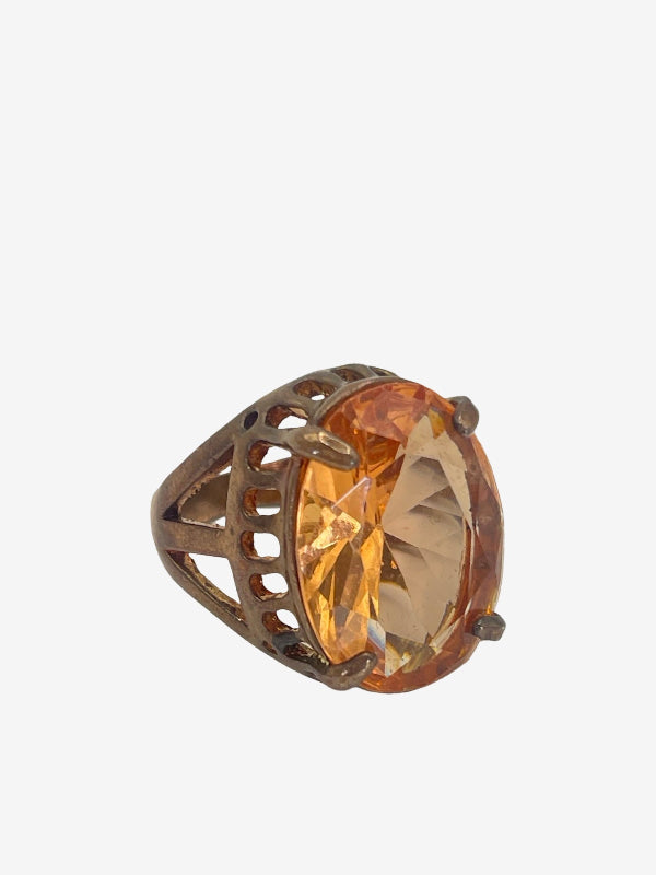 A ring featuring a large, oval-cut faceted amber-orange gemstone in an openwork bronze-toned metal setting, shown against a plain light gray background.