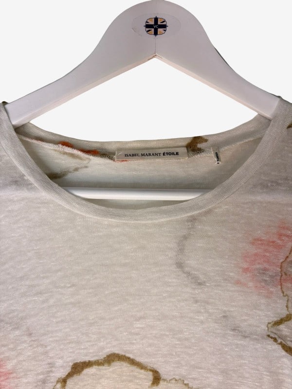 A close-up of a t-shirt hanging on a white hanger. The shirt is a light beige/off-white color with a watercolor-style print in brown, pink, and gold tones.