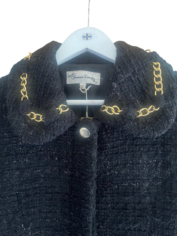 A close-up of a black textured tweed jacket hanging on a white hanger. The jacket features a large collar decorated with gold-tone chain embroidery and a silver-tone snap button at the center front.