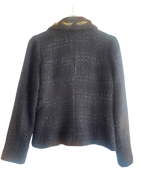 A dark black tweed cropped jacket with gold chain detailing along the collar, shown hanging against a white background.