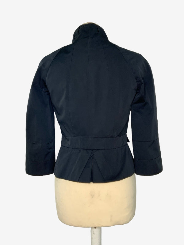 A black  jacket with a high collar and belted waist, shown from the back on a cream-colored dress form mannequin against a white background.