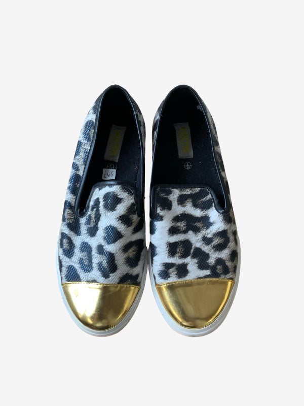A pair of slip-on sneakers with a black and white leopard print pattern and metallic gold toe caps, shown from a top-down view on a light gray background.