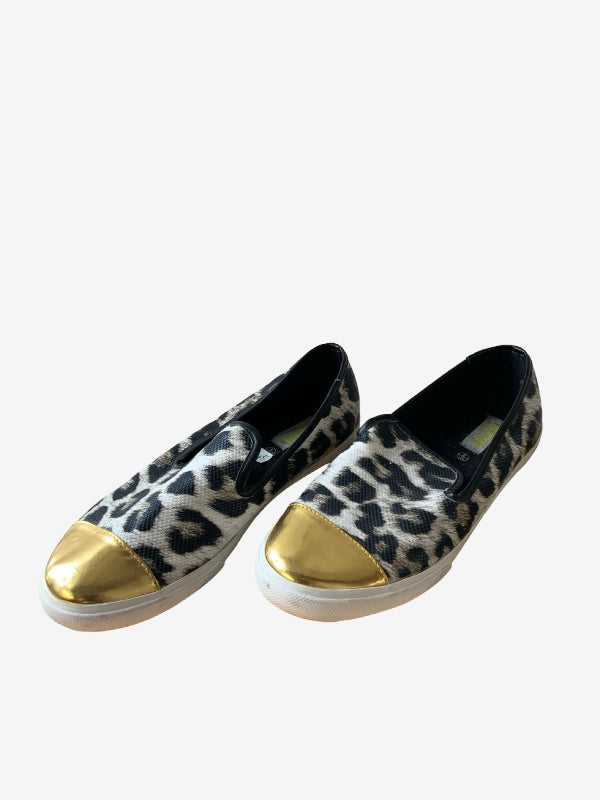 A pair of slip-on sneakers featuring a black and white leopard print pattern with metallic gold toe caps and white soles, shown against a plain light gray background.