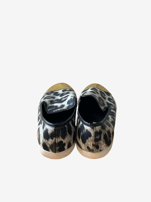A pair of slip-on shoes with a black and beige leopard print pattern and gold toe caps, shown from the back on a light gray background.