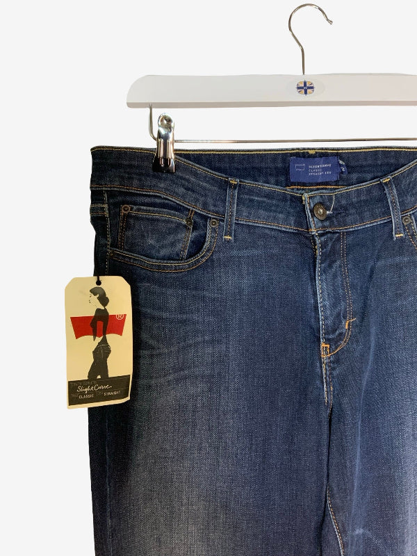 A pair of blue denim jeans hanging from a white hanger with a metal clip, shown against a plain white background. A paper brand tag hangs from the left side pocket.