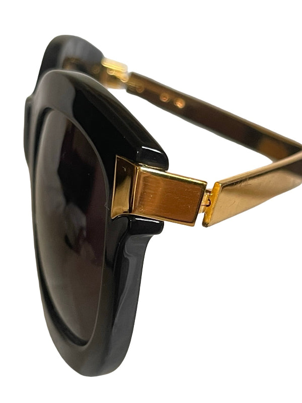 Close-up side view of oversized sunglasses with a glossy black frame and gold metal hinge details, set against a plain white background.