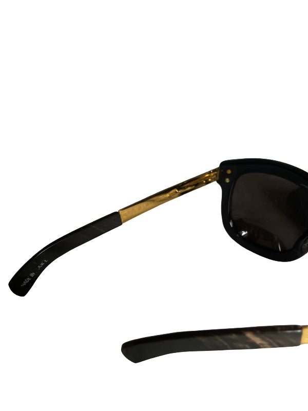 Close-up of sunglasses with black frames and dark lenses, featuring gold metal temples and a tortoiseshell-patterned lower temple arm, shown on a white background.