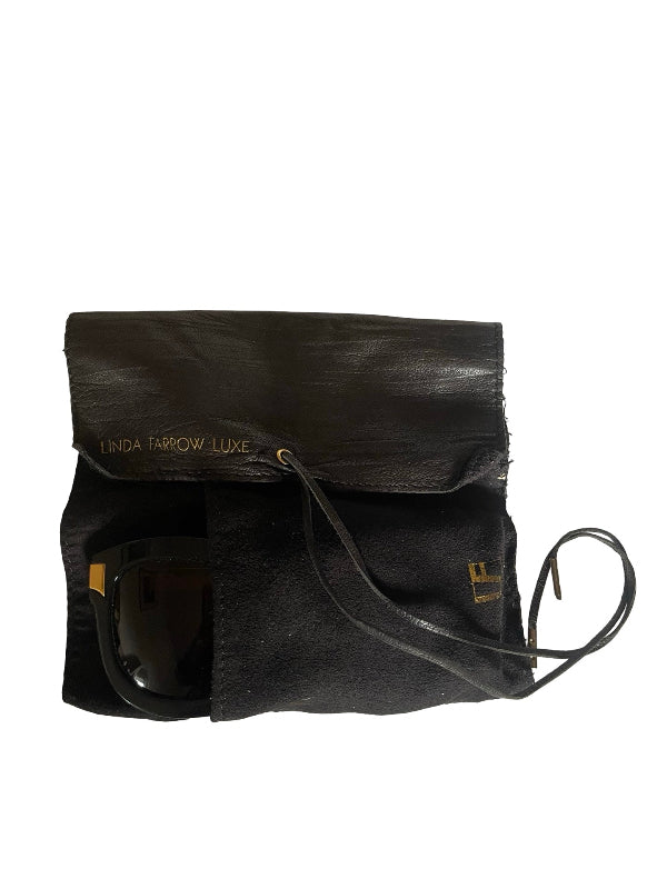 A black leather sunglasses pouch by Linda Farrell Luxe, featuring a flap closure with gold lettering, a front pocket, and a strap, shown on a plain white background with a pair of sunglasses inside.