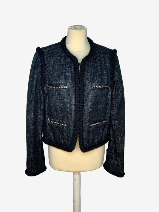 A charcoal tweed jacket with black braided trim and gold chain accents, displayed on a cream-colored mannequin torso against a light gray background.