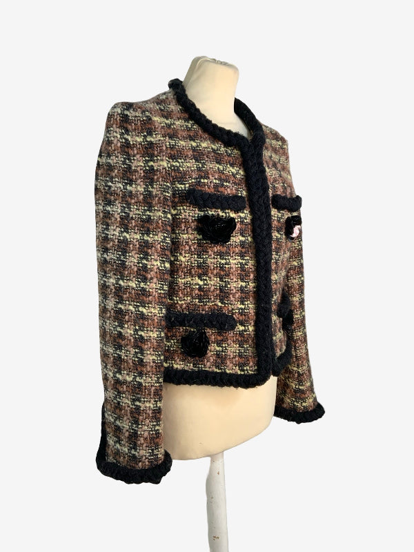 A cropped tweed jacket displayed on a cream mannequin torso against a light gray background. The jacket features a brown, beige, and black tweed pattern with black plaited trim along the collar, cuffs, pockets, and hem.