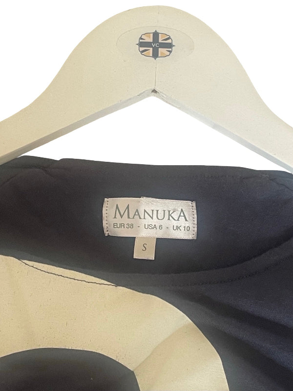 Close-up of a Manuka garment hanging on a cream-colored wooden hanger against a white background. The visible neck label reads MANUKA and shows size S.