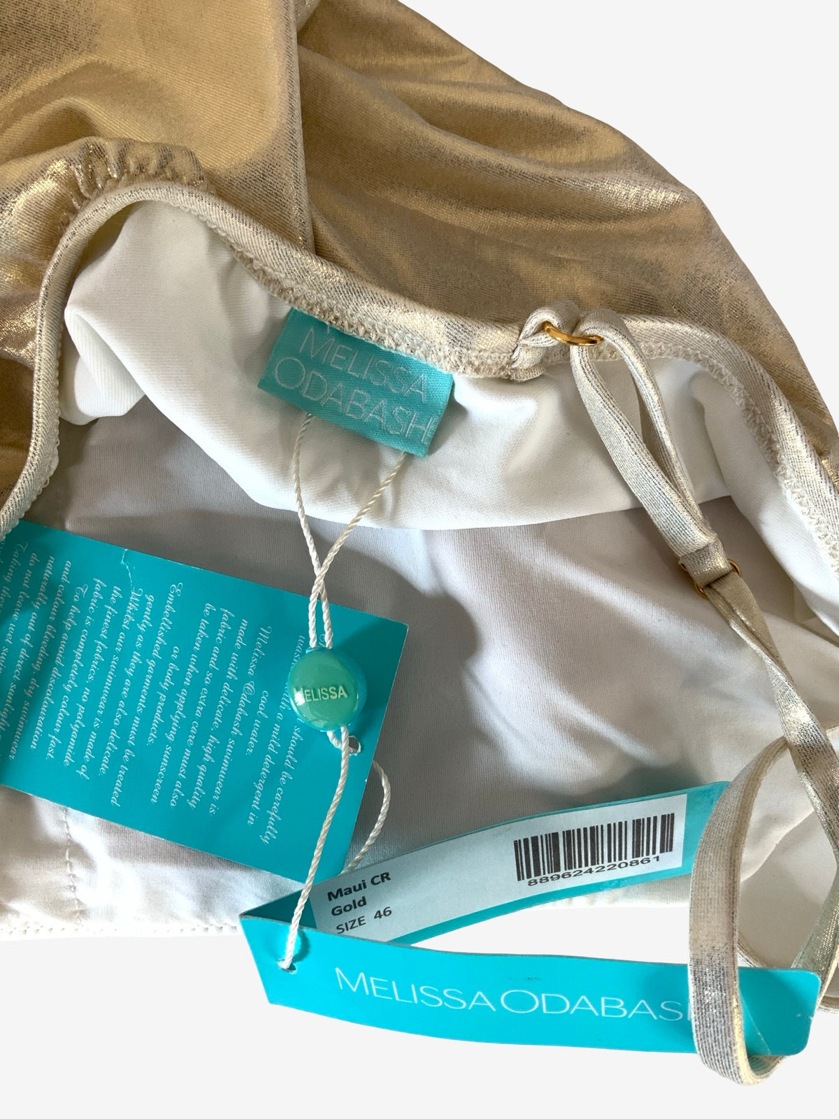 Close-up of a gold satin swimming costume with white lining, drawstring details, and multiple teal Melissa Odabash tags attached, photographed on a light neutral background.