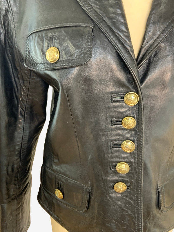 A close-up of a black leather jacket on a mannequin, featuring gold-tone decorative buttons on the flap chest pocket, a vertical row of large gold buttons on the front, and matching gold buttons on the lower flap pockets.