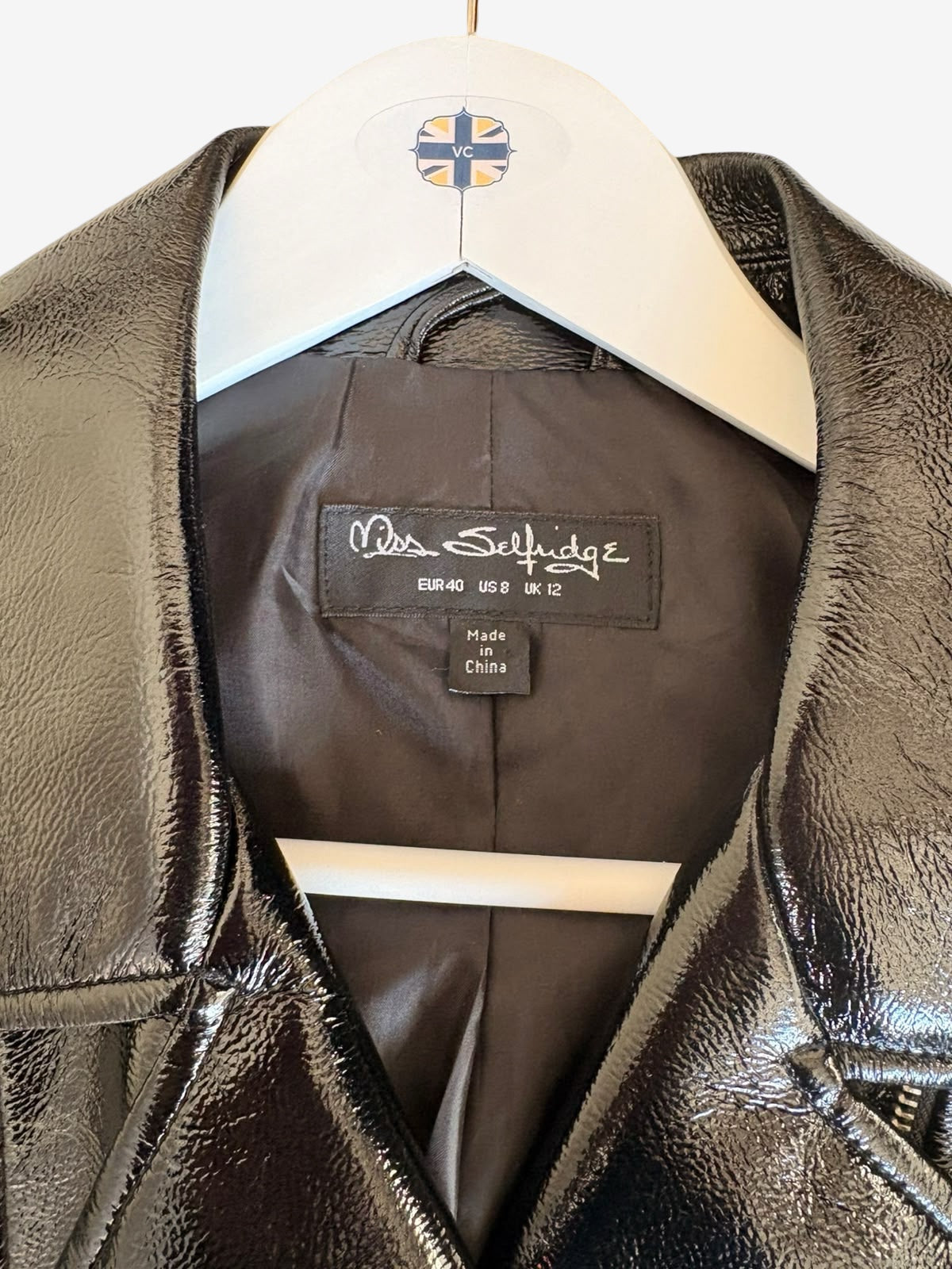 A close-up of a black leather jacket hanging on a white hanger with a visible neck label reading Miss Selfridge.
