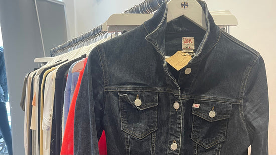 A dark wash denim jacket with a stand collar, displayed on a white hanger on a clothing rack alongside other garments.